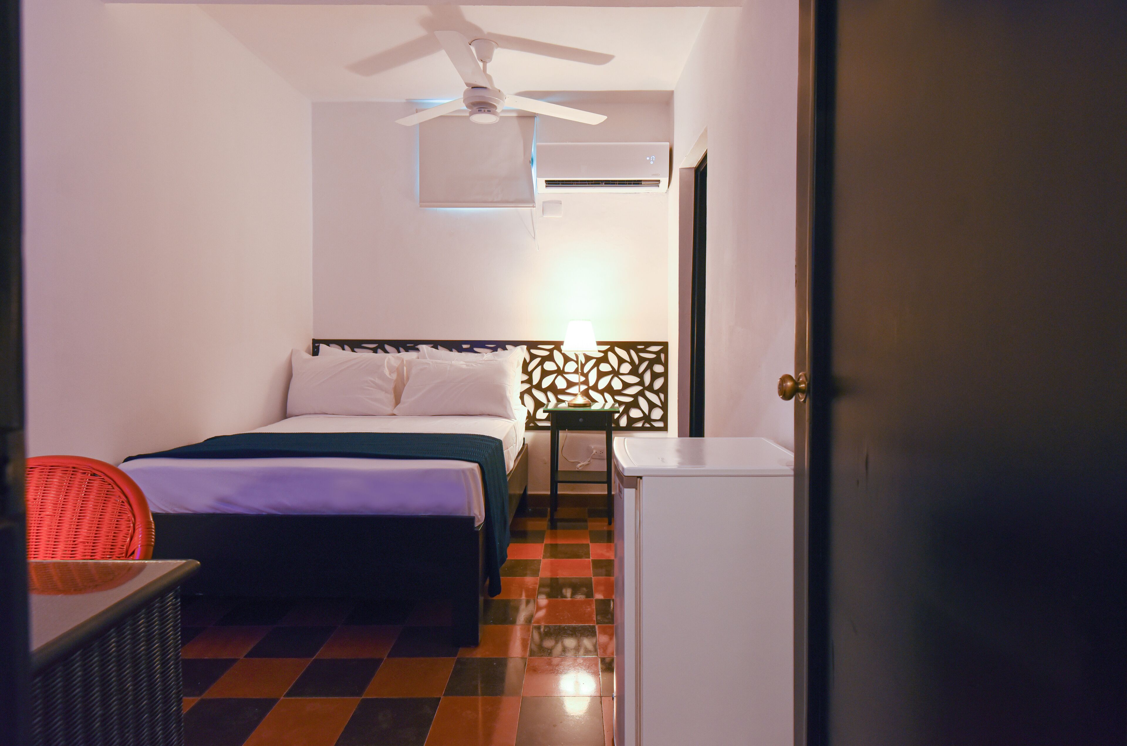 A hotel room with a bed, a chair, a desk, a ceiling fan, an air conditioner, and a door.