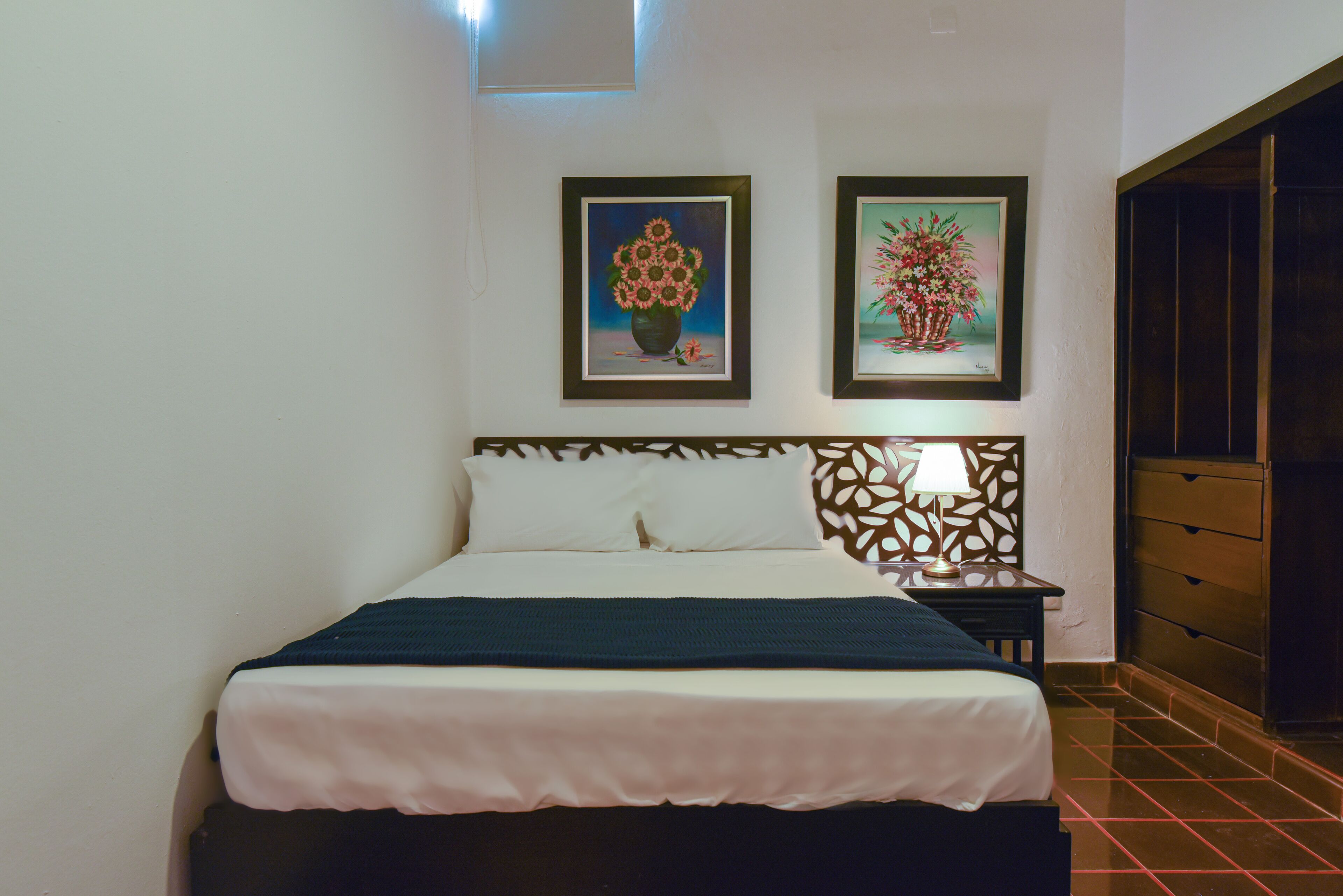 A neatly made bed with a white and teal bedspread, a wooden headboard, and two framed floral paintings on the wall.