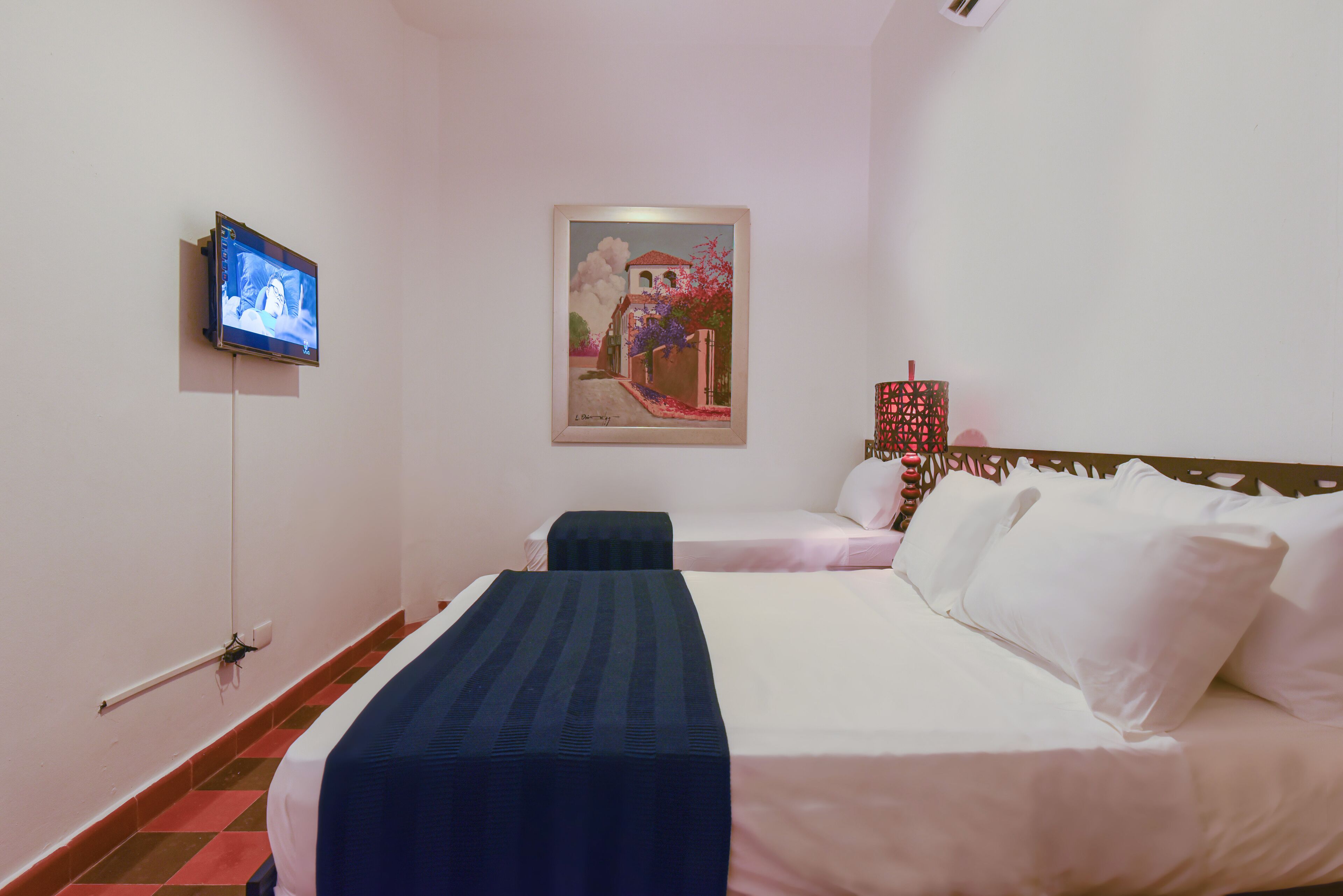 A hotel room with two beds, a TV mounted on the wall, a painting above the beds, and a patterned lamp on the nightstand.