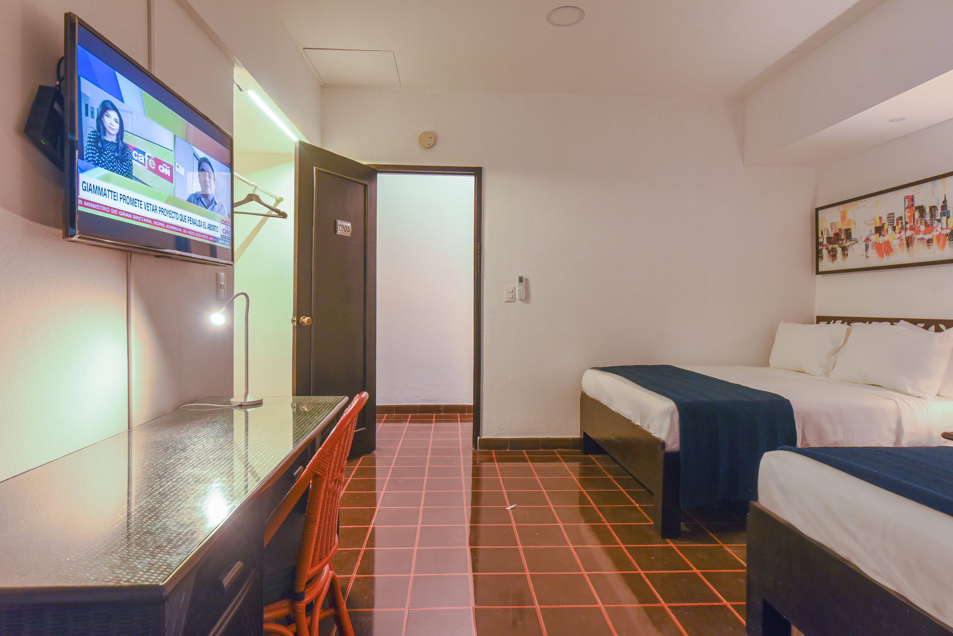 Habitación de hotel con televisor en la pared, escritorio, dos camas y piso de baldosas.