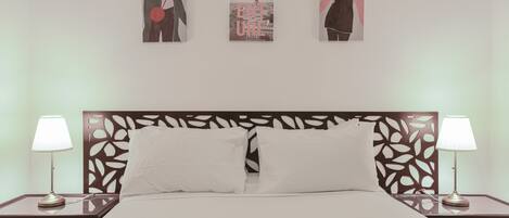 Een netjes opgemaakt bed met witte lakens, twee nachtkastjes met lampen en drie ingelijste kunstwerken aan de muur.