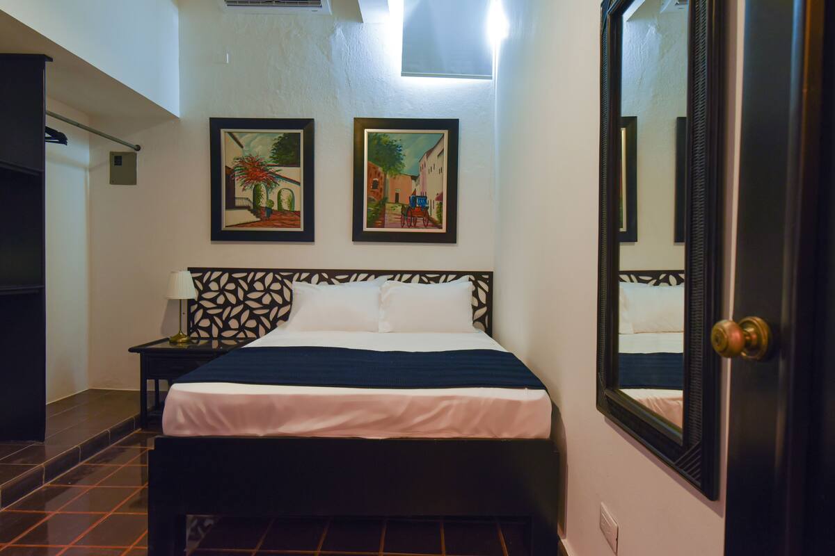 Ein Hotelzimmer mit einem Bett, einem Nachttisch, einer Lampe, zwei gerahmten Bildern an der Wand und einem großen Spiegel.