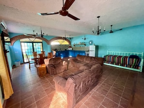 Uma sala de estar espaçosa com um sofá marrom grande, uma área de jantar com cadeiras de madeira, uma cozinha com armários azuis e um ventilador de teto.