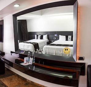 Habitación de hotel con dos camas, un escritorio con espejo, y una bandeja con agua embotellada y pañuelos.