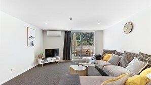 A modern living room with a grey sofa, a coffee table, a TV on a white stand, and a large window with a view of greenery. - Central Mountain View Paradise (Queenstown)