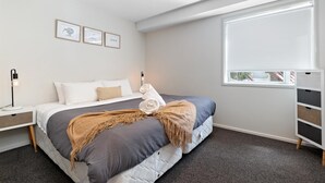 A modern bedroom with a large bed, a bedside table, a nightstand, a window with blinds, and framed artwork on the wall. - Central Mountain View Paradise (Queenstown)