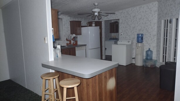 A compact kitchen with a central island, wooden stools, a refrigerator, a washing machine, and a ceiling fan. - Excellent Recreational Farm Minutes from DineyWorld (Frostproof)