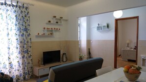 A living room with a sofa, TV, and a dining area with a table and chairs. There are shelves with decorative items and a plant on the wall.