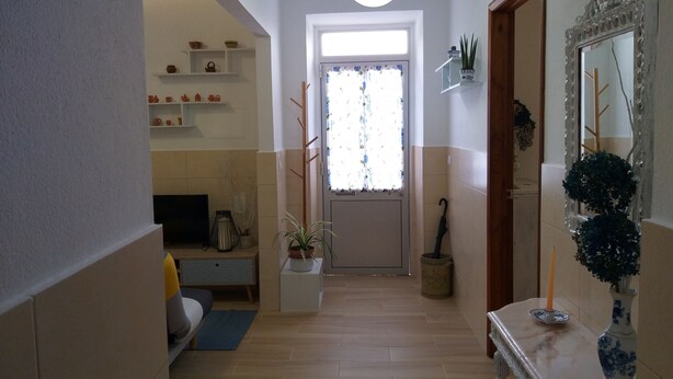 A hallway with a wooden floor, a white door with a window, a mirror with a decorative frame, and a small table with a vase.