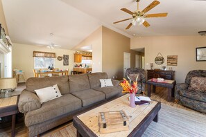 A living room with a sofa, coffee table, and chairs. There is a dining area with a table and chairs. A ceiling fan is visible. - Lazy Squirrel - Cozy Family House. Game Room. Mountains. 10 min River (Kernville)