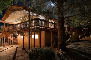 A two-story wooden cabin with a balcony, surrounded by trees and a dirt ground. - Lazy Squirrel - Cozy Family House. Game Room. Mountains. 10 min River (Kernville)