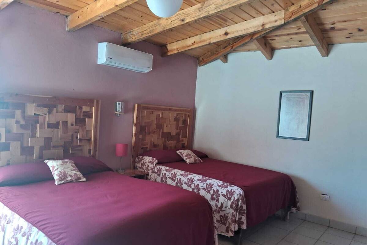 A room with two beds, wooden ceiling beams, an air conditioning unit, a wooden headboard, and a framed picture on the wall.