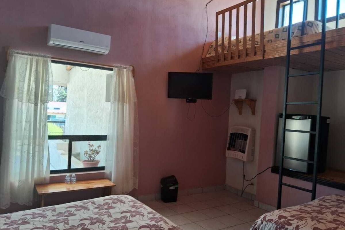 A dormitory room with bunk beds, a window with curtains, an air conditioner, a television, and a small table.