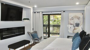 A modern bedroom with a large bed, a flat-screen TV, a bench, and a balcony with a view of trees. - Luxury Mountain Suite - Ski/Golf/Hike/Play (Killington)
