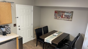 Een kleine keuken met een vaatwasser, een koffiezetapparaat en een stapel blikjes. Er staat een eettafel met stoelen en er hangt een schilderij aan de muur.
