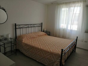 Un lit simple avec une couvre-lit à motifs, une table de chevet avec une lampe et une fenêtre ornée de rideaux légers.