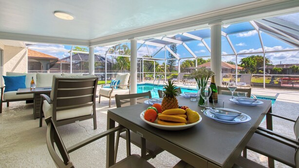 A covered outdoor dining area with a pool view, a table set with fruit and a bottle, and chairs around. - VRCC Villa San Miguel (Cape Coral)