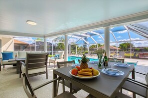 A covered outdoor dining area with a pool view, a table set with fruit and a bottle, and chairs around.