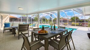 A sunroom with a pool view, featuring a table with fruit, a bottle, and glasses. - VRCC Villa San Miguel (Cape Coral)