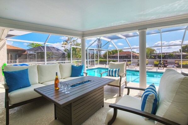 A sunlit outdoor seating area with a pool, white cushioned chairs, and a wooden table.