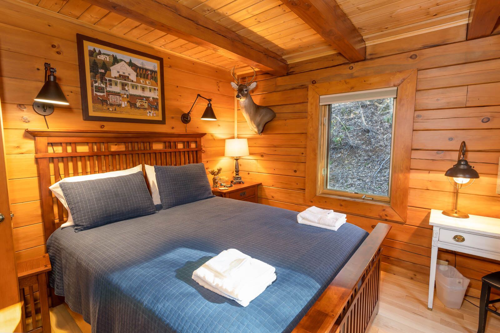 A wooden cabin room with a bed, a nightstand, a lamp, a window, and a framed picture on the wall.