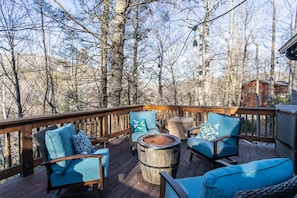 A wooden deck with outdoor seating, a fire pit, and a view of a forested area.