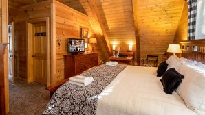 A cozy attic bedroom with a wooden ceiling, a bed with white linens, a dresser, a television, and a desk with a lamp. - Cozy Cabin Tucked Away in Wooded Property (Boone)