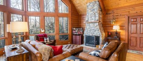 A cozy log cabin interior with a stone fireplace, leather sofas, and large windows offering a view of the forest.