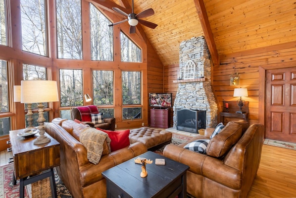A cozy log cabin interior with a stone fireplace, leather sofas, and large windows offering a view of the forest.
