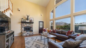 A spacious living room with a stone fireplace, a leather sofa, and large windows offering a mountain view. - Epic Mountain View. Luxury House on a Hill. River 10 min (Kernville)
