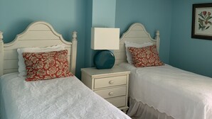 Quarto com duas camas, cada uma com cabeceira branca e almofadas floridas vermelhas. Há uma mesa de cabeceira branca com abajur azul entre as camas. As paredes são pintadas de verde-água, e há um quadro na parede.