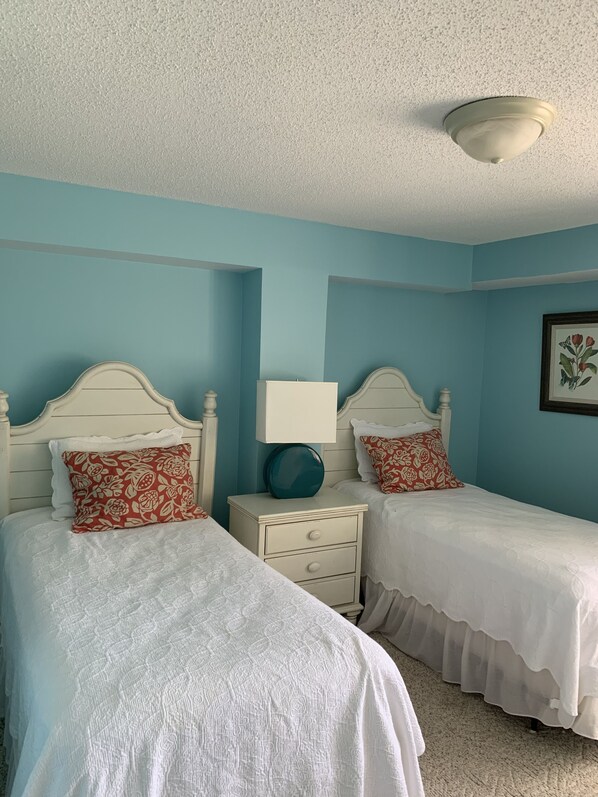 A bedroom with two beds, each with a white headboard and red floral pillows. There is a white nightstand with a blue lamp between the beds. The walls are painted teal, and there is a framed picture on the wall.