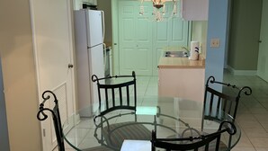 Uma sala de jantar com mesa e cadeiras de vidro, um lustre e uma cozinha ao fundo.