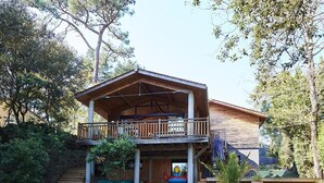 Une maison en bois à deux étages, dotée d’un balcon, entourée d’arbres et bordée d’un terrain sablonneux.