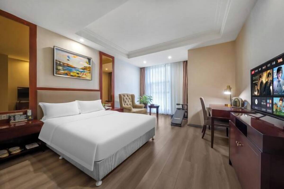 A hotel room with a large bed, a desk, a chair, a TV, and a painting on the wall.
