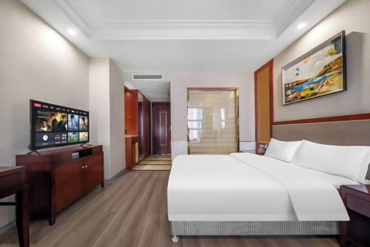 A hotel room with a bed, a TV on a wooden cabinet, a painting on the wall, and a door leading to another room.
