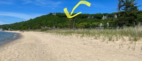 A sandy beach with a clear sky, a grassy area, and a hill with trees and buildings in the background.