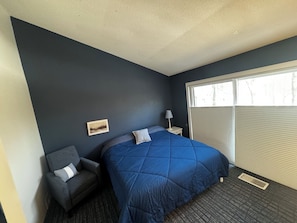 Un dormitorio con pared azul oscuro, una cama con colcha azul, un sillón gris, una mesita de noche con lámpara y una ventana con persianas.