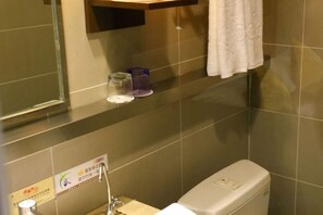 Casa de banho com sanita, lavatório e espelho. Uma toalha está pendurada na parede e há uma pequena estante com um copo e um objeto roxo.