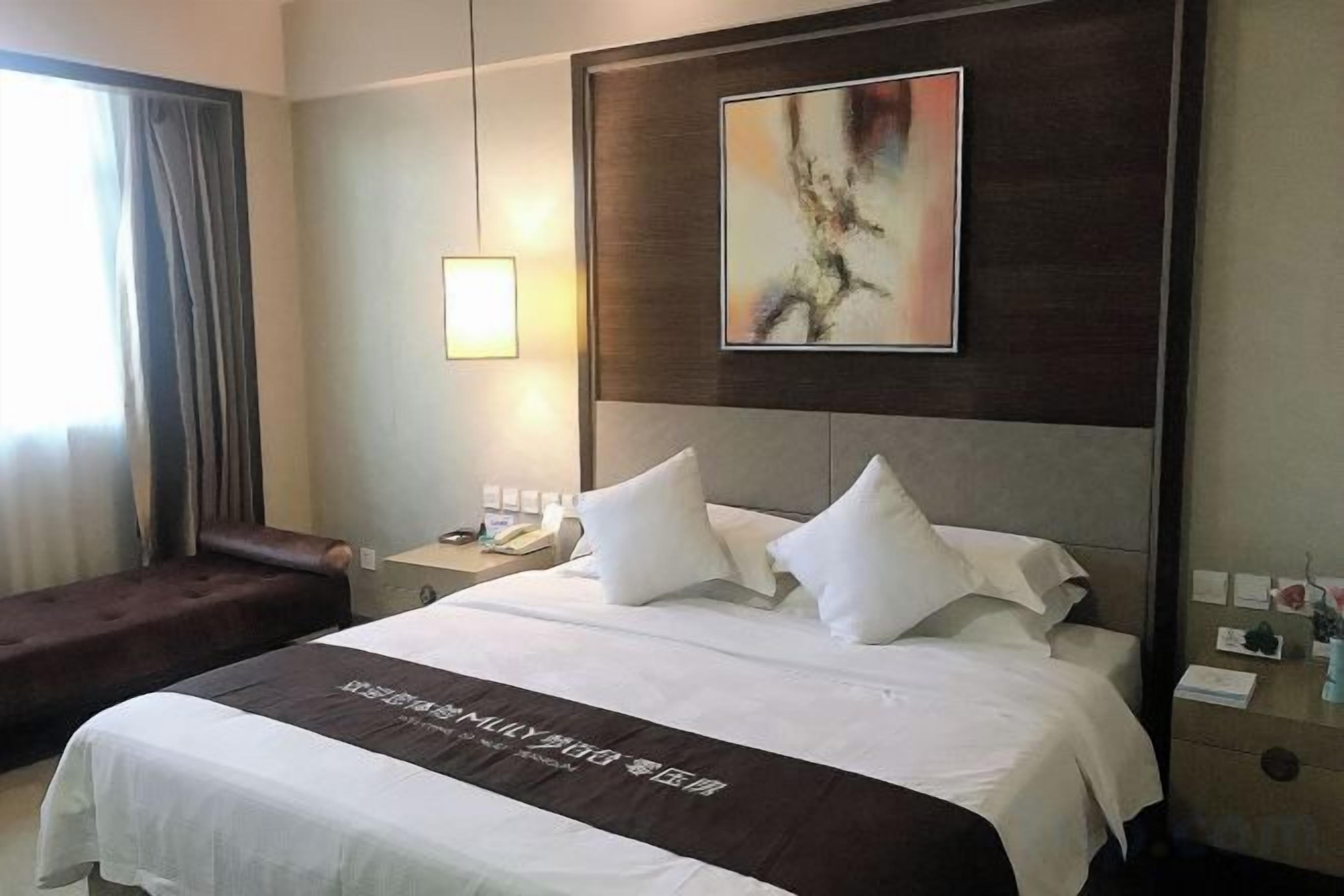 A hotel room with a large bed, a bedside table, a lamp, a painting on the wall, and a window with curtains.