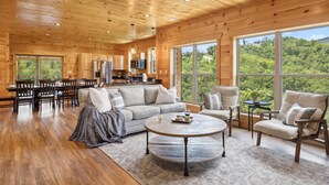 A spacious living room with a wooden ceiling, large windows offering a view of the forest, a dining area with a wooden table and chairs, and a kitchen with modern appliances. - Ridgeline Retreat w/ Heated Pool, Hot Tub + Views | Sleeps 12 (Sevierville)