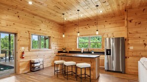 A wooden cabin interior with a kitchen area featuring a stainless steel refrigerator, wooden bar stools, and large windows. - Ridgeline Retreat w/ Heated Pool, Hot Tub + Views | Sleeps 12 (Sevierville)