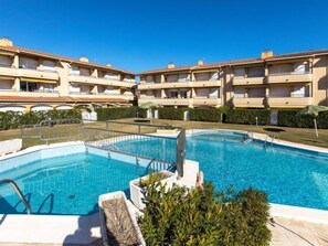 A swimming pool with a clear blue water, surrounded by a white pool deck and a metal safety railing. In the background, there are multi-story buildings with balconies and a well-maintained hedge.