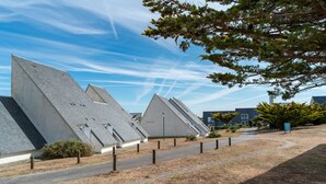 A row of modern, triangular buildings with sloped roofs, a paved walkway, and a clear blue sky. - 200m de la Plage | Apt Cosy pour 3 + Terrasse (Guidel)