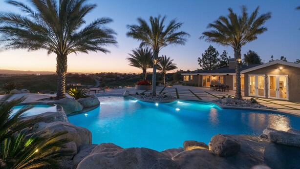 A swimming pool surrounded by large rocks and palm trees, with a house in the background. - Wine Country Retreat • Pool, Spa, Views (Temecula)