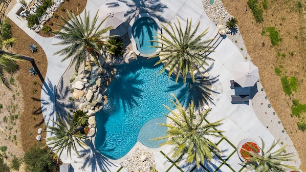 An aerial view of a swimming pool surrounded by palm trees and rocks, with a white tiled deck and umbrellas.