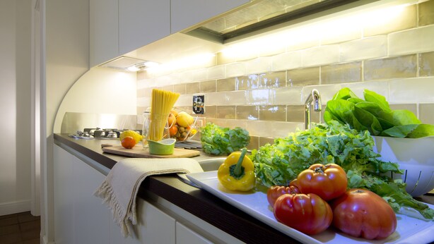 Une cuisine avec un plan de travail blanc, un évier et divers légumes, comme des tomates et des poivrons.