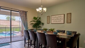 A dining area with a table set for six, surrounded by chairs, a chandelier, and a view of a swimming pool through large windows. - Epic 9BR Game House - Arcade, PS5, Pool & Hot Tub (Davenport)