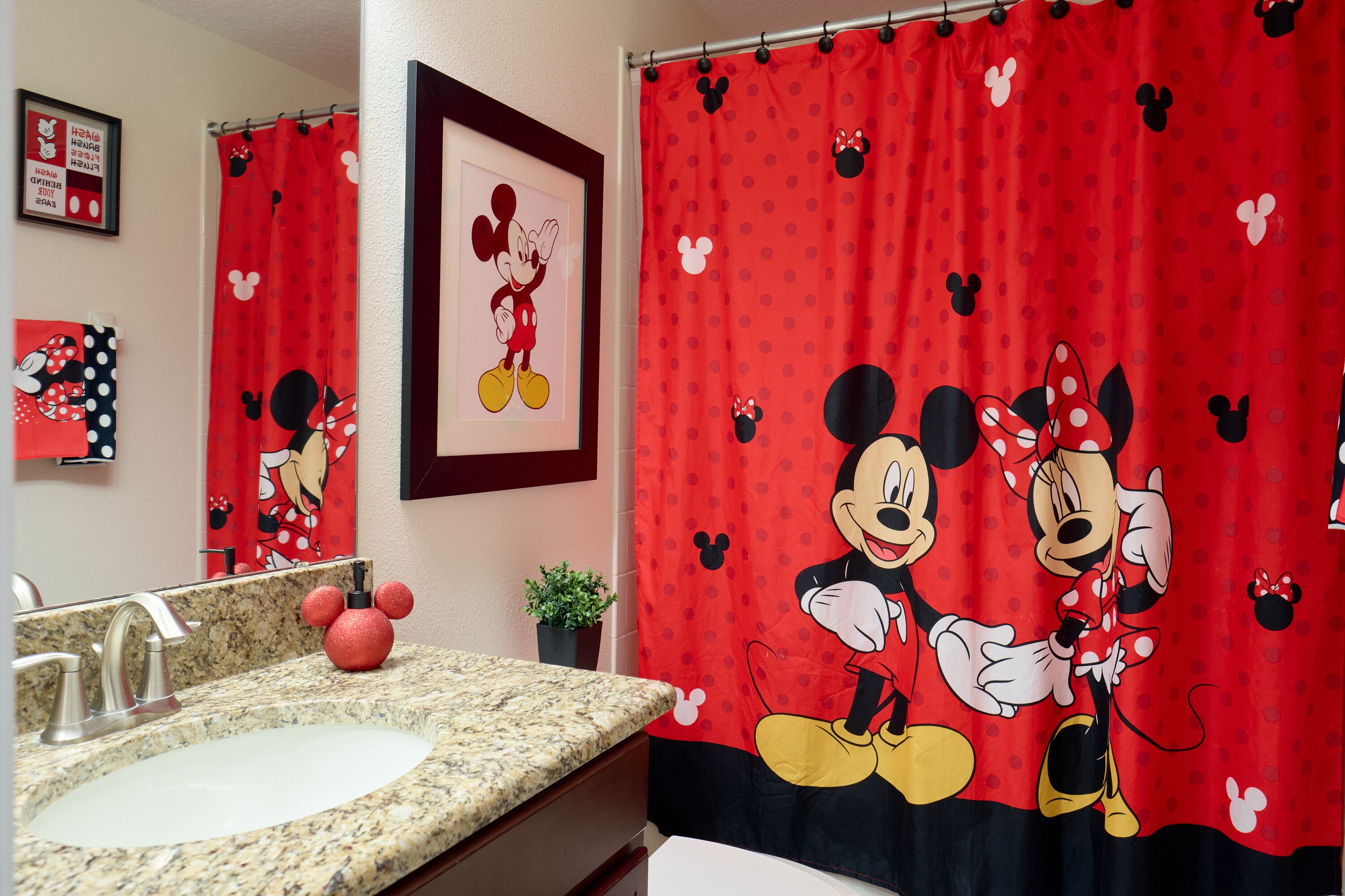 Et badeværelse med et bruseforhæng med Mickey Mouse tema, en vask med granitbordplade og et indrammet billede af Mickey Mouse på væggen.