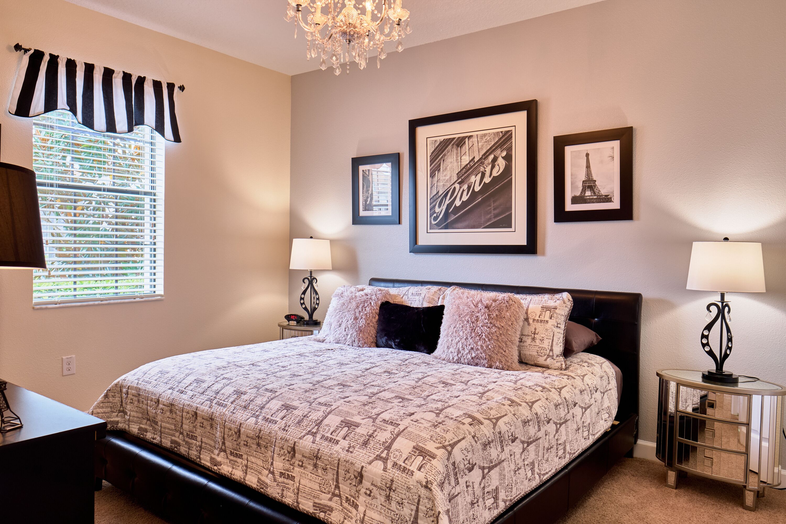 A bedroom with a large bed, two bedside tables with lamps, a chandelier, and three framed pictures on the wall.